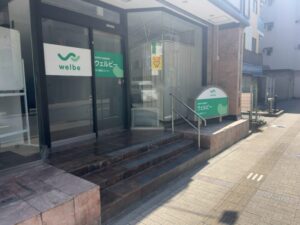 ウェルビー本八幡南口センターの外観。入口前の階段と手すり、ガラス面のロゴ掲示、右手に屋外看板が見える(2026年3月5日筆者撮影)