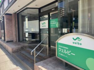 ウェルビー本八幡南口センターの入口付近。ガラス面に「welbe」ロゴと「ウェルビー 本八幡南口センター」の掲示、建物前に看板が設置されている様子（2026年3月5日撮影）
