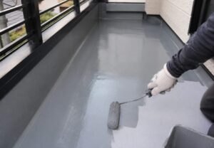 ベランダの床面にグレーの防水塗料をローラーで塗布している防水工事中の写真